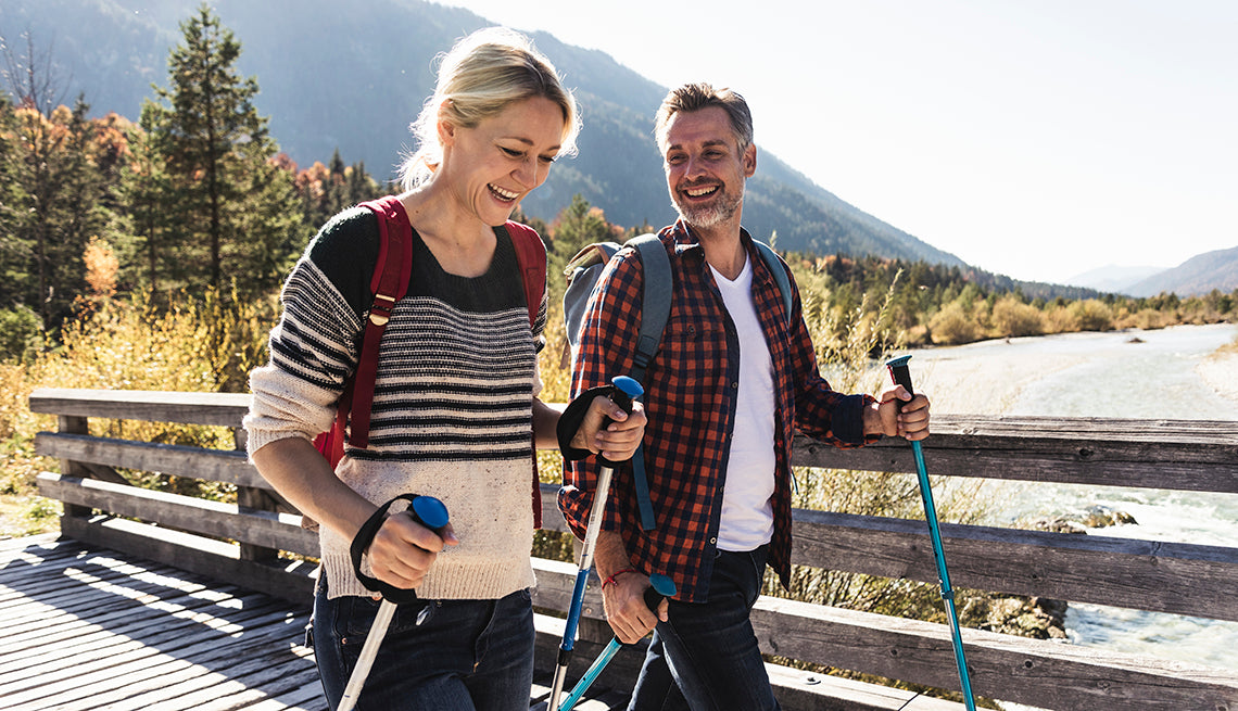 Two hikers with backpacks and walking sticks on a wooden bridge with mountains in the background